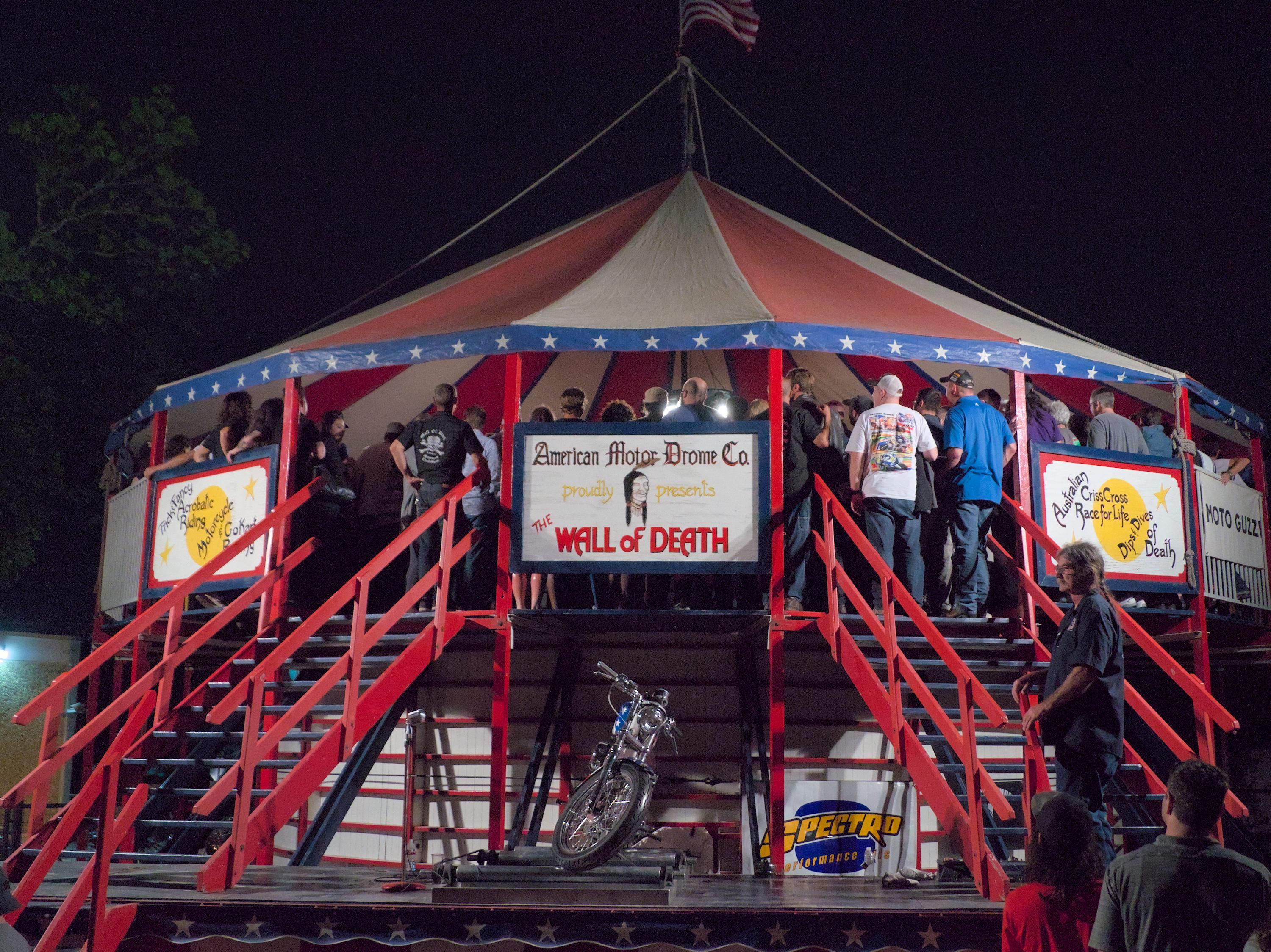 A large elevated tent with a group of people down inside watching the wall of death show where motorcycles ride along the inner walls doing stunts